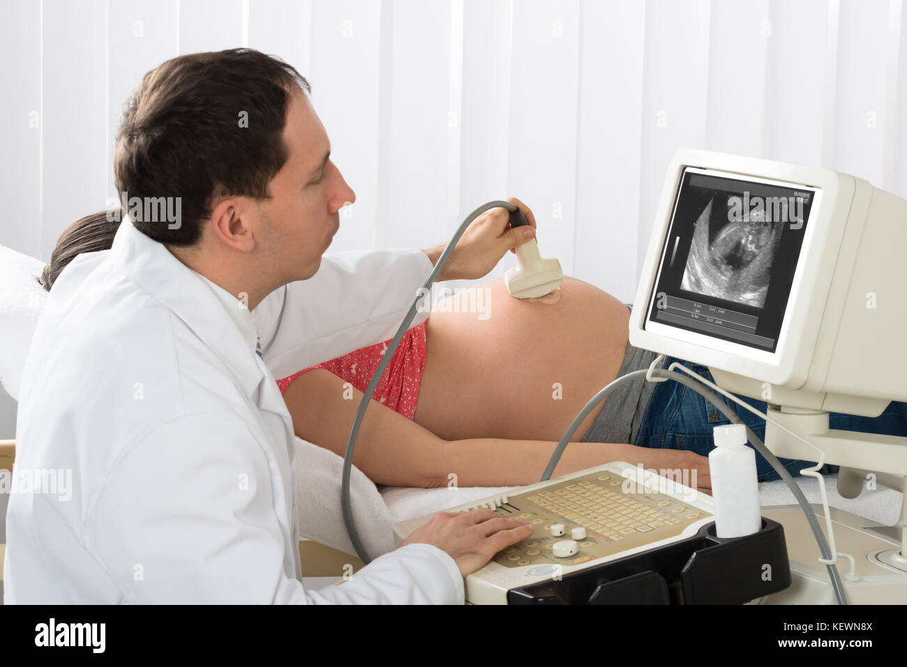Felice medico maschio muovendo il trasduttore di ultrasuoni sulla donna incinta la pancia mentre guardando lo schermo Foto Stock