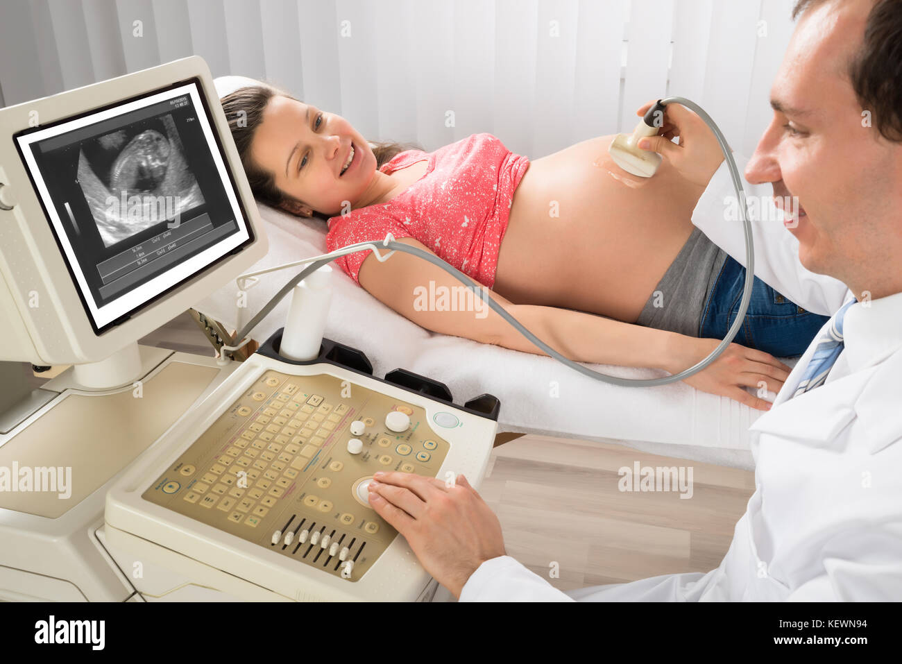 Felice medico maschio muovendo il trasduttore di ultrasuoni sulla donna incinta la pancia mentre guardando lo schermo Foto Stock