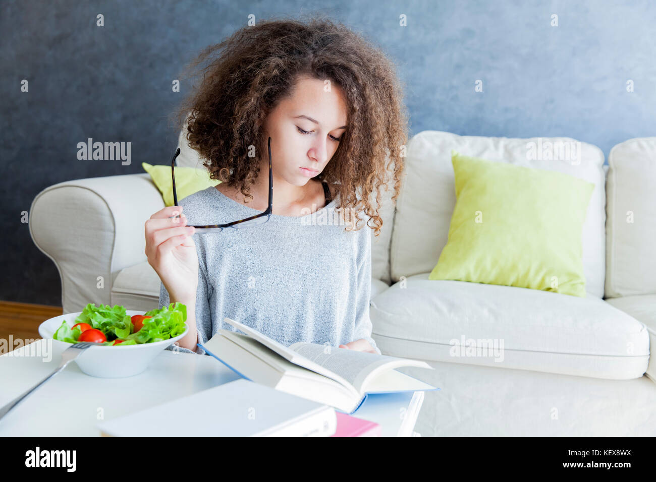 Capelli ricci teen ragazza libro di lettura e mangiare insalata in camera Foto Stock