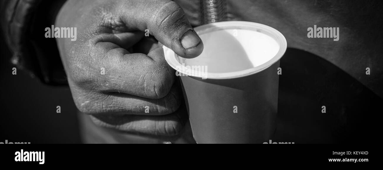 Sporco mendicante mano che regge una tazza di plastica. Povero mendicante di denaro sulla strada vicino. Foto Stock