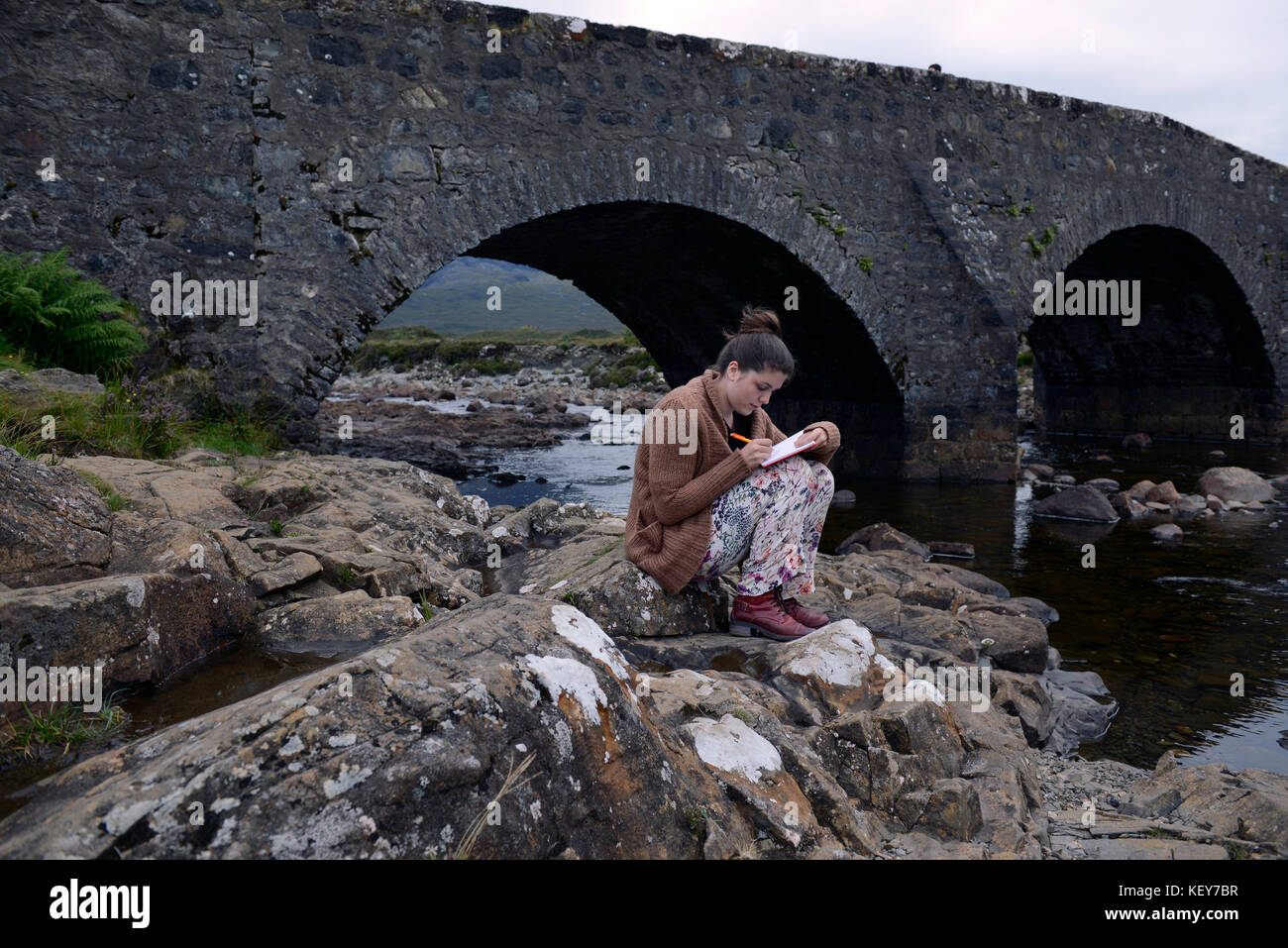La diciassettenne scrive nel suo diario sul vecchio ponte di pietra sul fiume Slichagan, Isola di Skye, Scozia Foto Stock