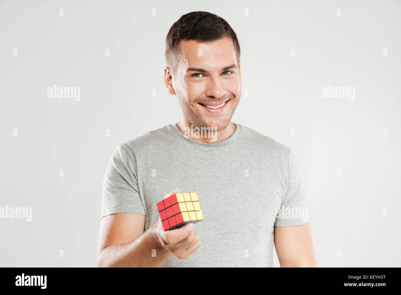 Immagine del giovane uomo felice vestito in grigio t-shirt isolate su parete grigia. Guardando la telecamera tenendo premuto il cubo di Rubik. Foto Stock