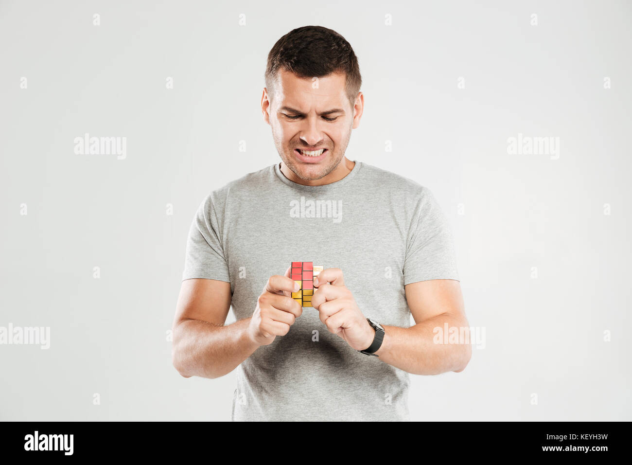 Foto di giovani confusi uomo vestito in grigio t-shirt isolate su parete grigia. Guardando a parte provare a risolvere il cubo di Rubik. Foto Stock