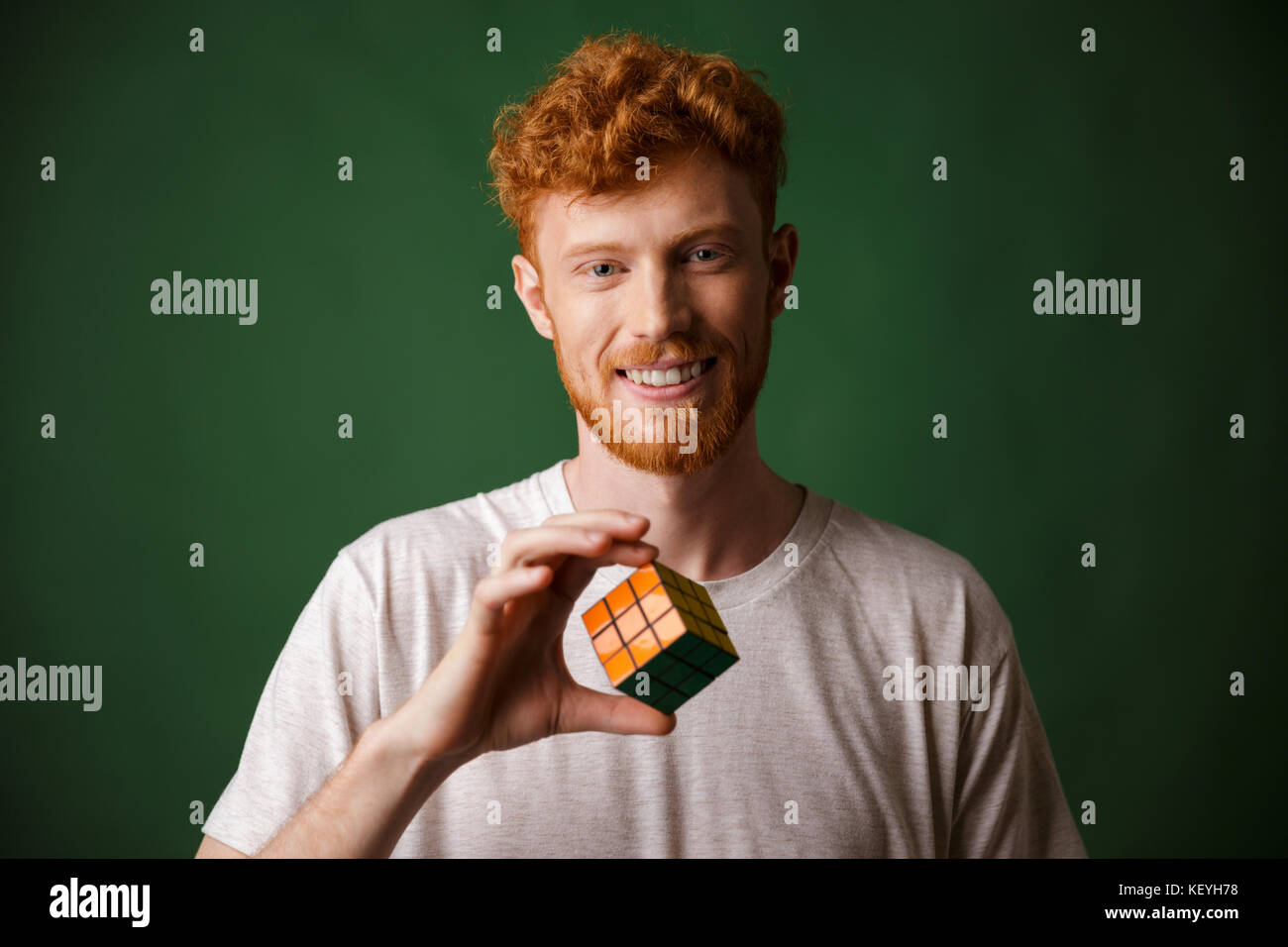 Close-up shot di giovani readhead sorridente uomo barbuto, tenendo il cubo di Rubik, su sfondo verde Foto Stock