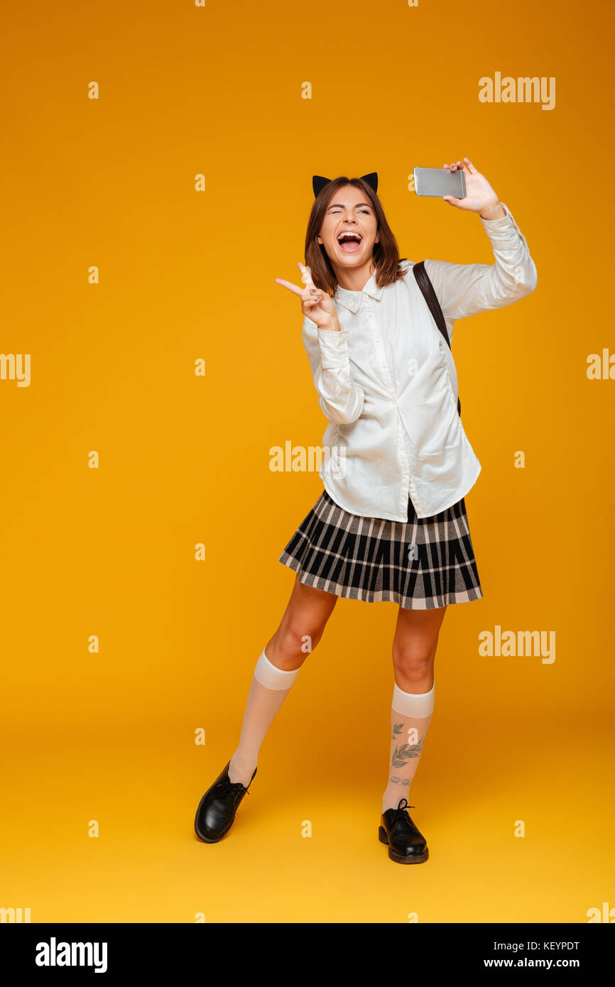 A piena lunghezza Ritratto di un felice teenage schoolgirl in uniforme con zaino prendendo un selfie mentre in piedi e baluginare isolate su sfondo arancione Foto Stock