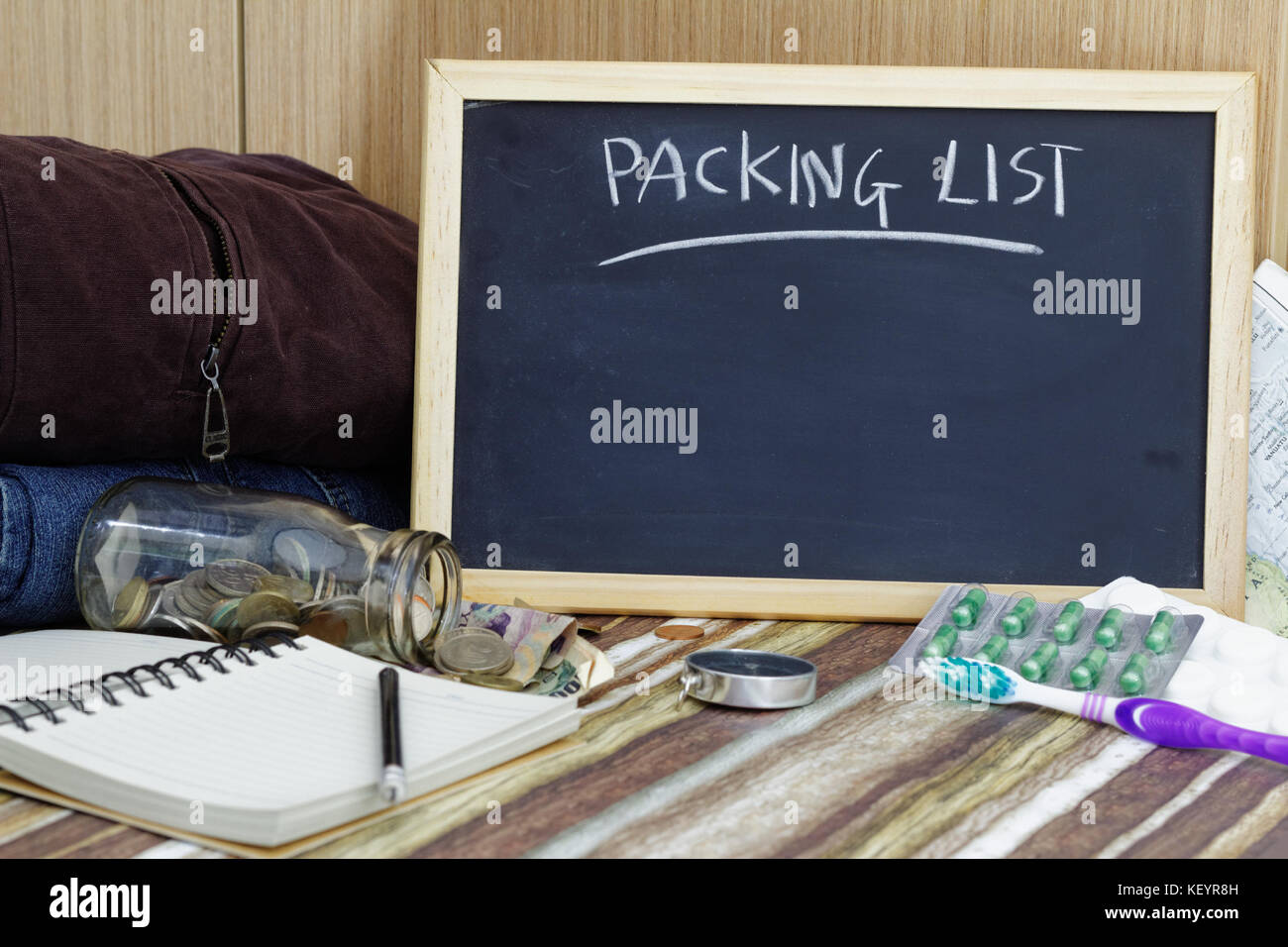 Concetto di viaggio. imballaggio elenco sulla lavagna nera con vestiti,medicina,bussola,spazzolino da denti,matita,vuoto note book e denaro fuoriuscita del vasetto di vetro. Foto Stock