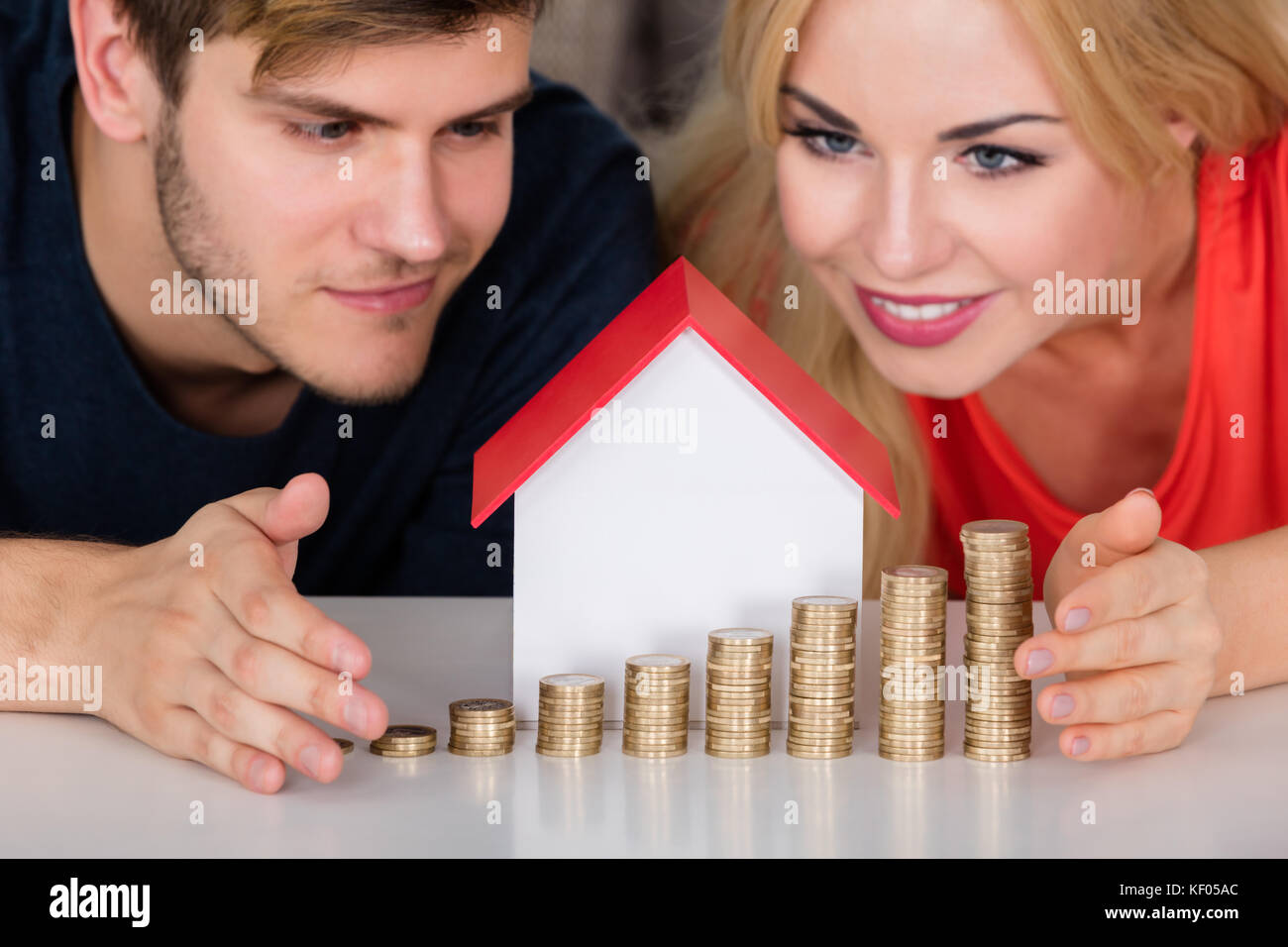 Giovane coppia sorridente proteggere casa modello con monete impilate sulla scrivania. Affitto Immobili concetto di crescita Foto Stock