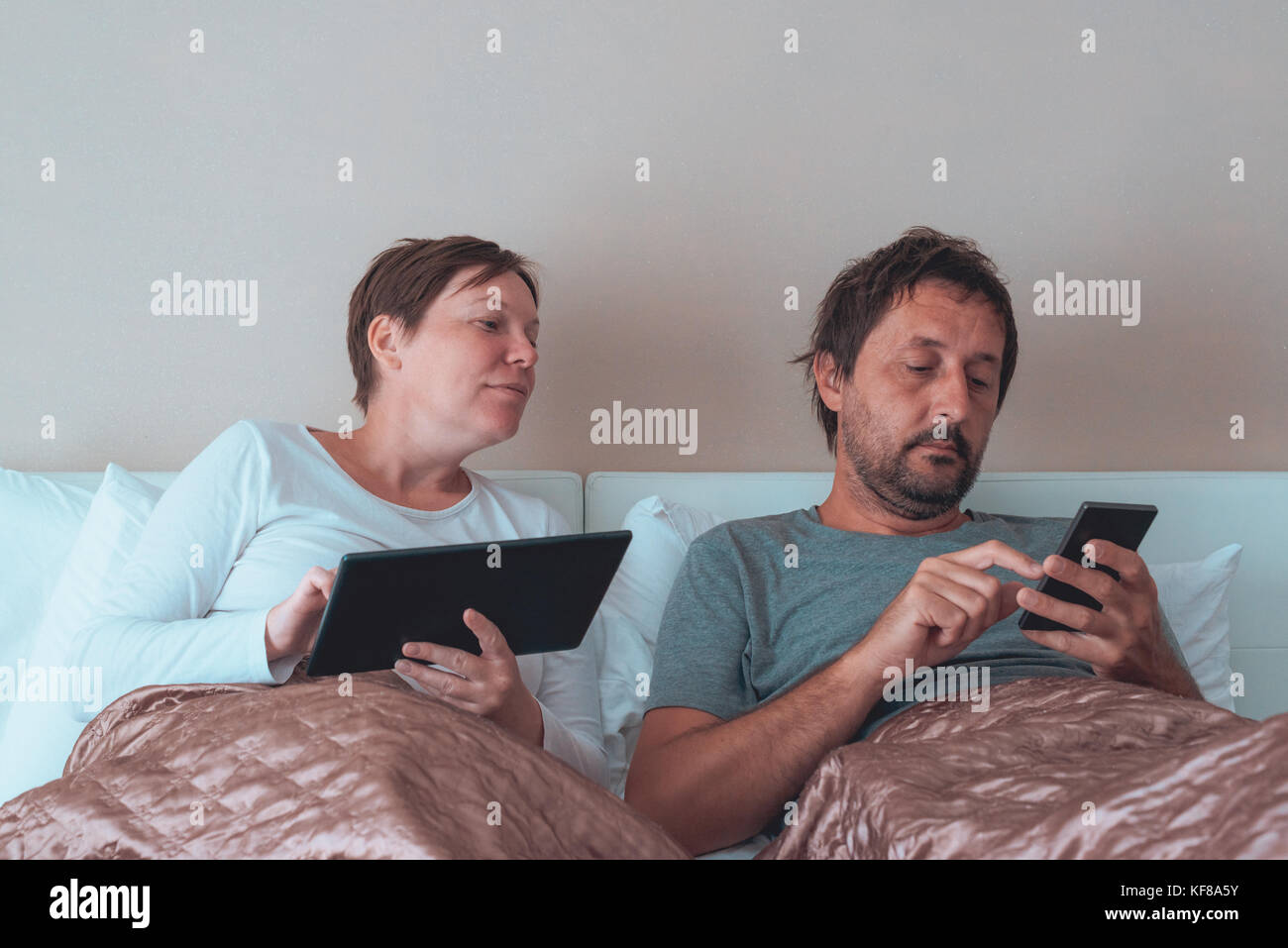 Annoiato giovane, il marito e la moglie in camera da letto utilizzando dispositivi elettronici personali Foto Stock