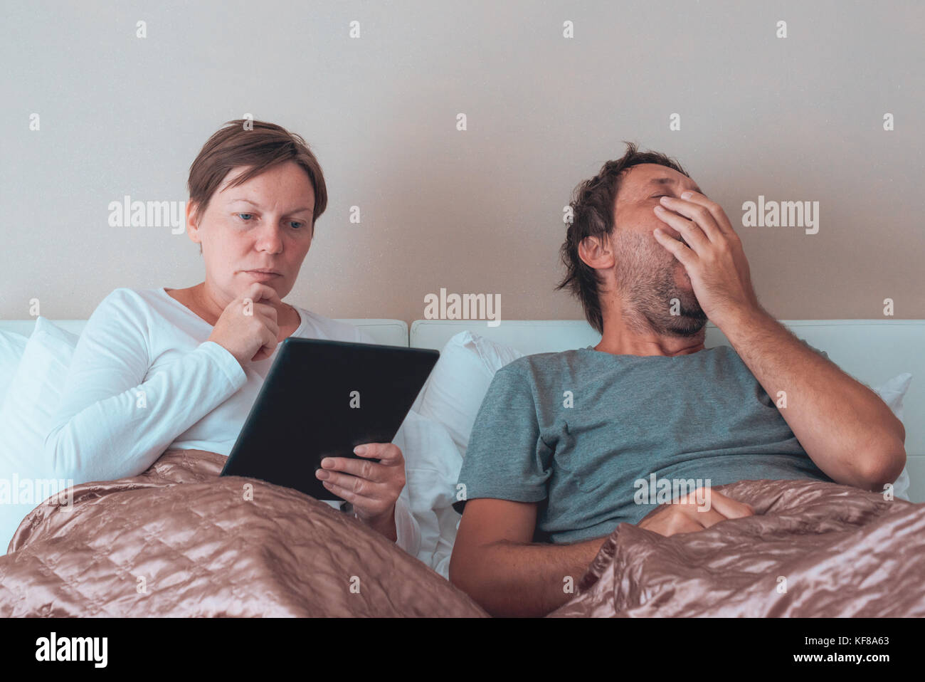 Annoiato giovane, il marito e la moglie in camera da letto utilizzando dispositivi elettronici personali Foto Stock