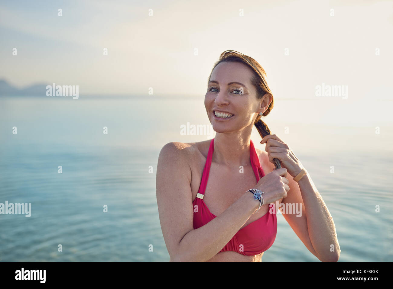 Sorridenti donna di mezza età mantenendo i suoi lunghi capelli in piedi con la schiena verso l'oceano e bagliore del sole al mare come ella gode di un giorno Foto Stock