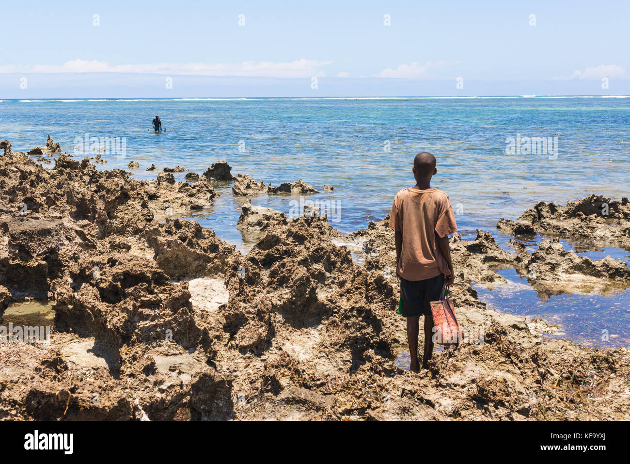 Giovane ragazzo africano che porta con sé una vecchia borsa della spesa e si trova su una costa rocciosa in attesa di un pescatore di lancia che stava tornando dal mare. Mare blu e. Foto Stock