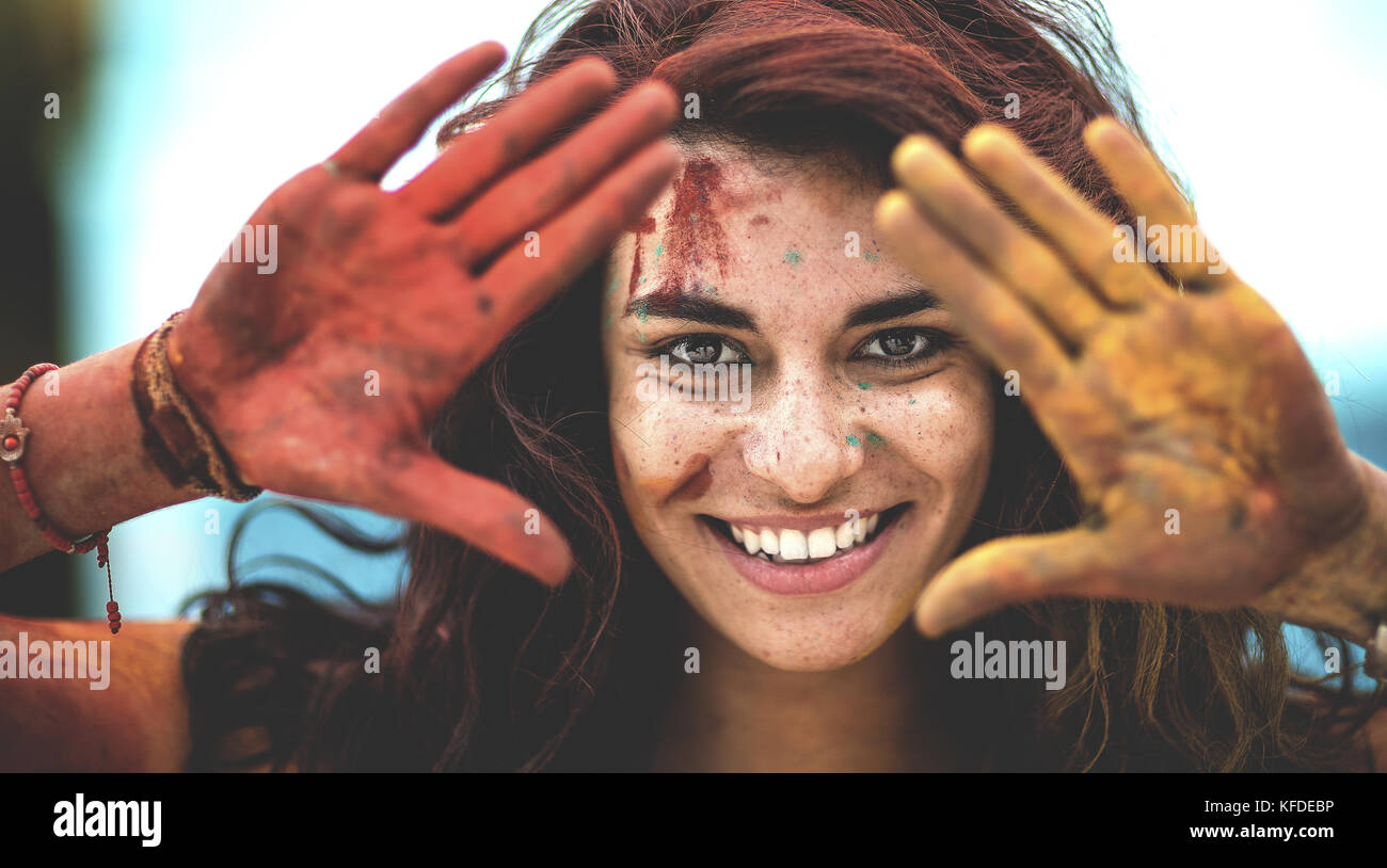 Giovane donna tenendo coperto di vernice mani fino al suo volto, una rossa e una gialla, sorridente, che incorniciano il suo volto. Foto Stock