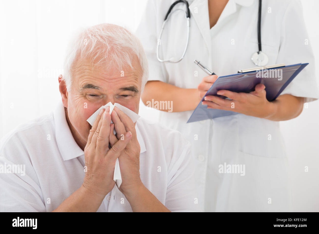 Senior uomo infettati con il freddo soffia il naso mentre medico iscritto negli appunti in clinica Foto Stock
