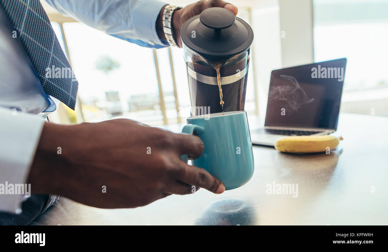 Imprenditore versando il caffè caldo in una tazza al tavolo della colazione con un computer portatile dal lato. Foto Stock