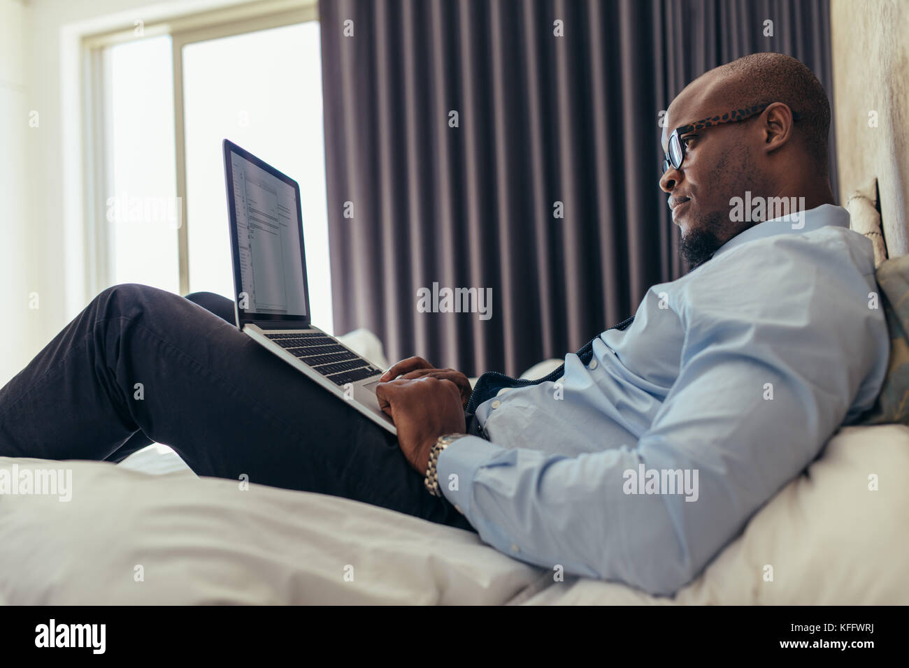Uomo in abiti formali lavorando su laptop mentre giaceva a letto. Imprenditore lavora su computer portatile dalla sua camera d'hotel. Foto Stock