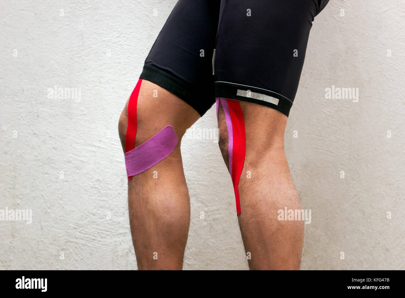 La foto in orizzontale di un ciclista maschio con nastro kinesio sulle ginocchia in piedi di fronte a uno sfondo bianco verso sinistra Foto Stock