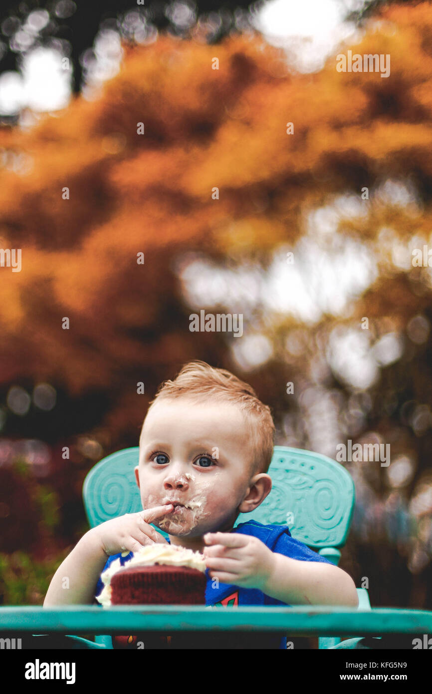 Ampia eyed baby boy di mangiare la torta nel suo seggiolone blu. alberi sfocata in background. Foto Stock