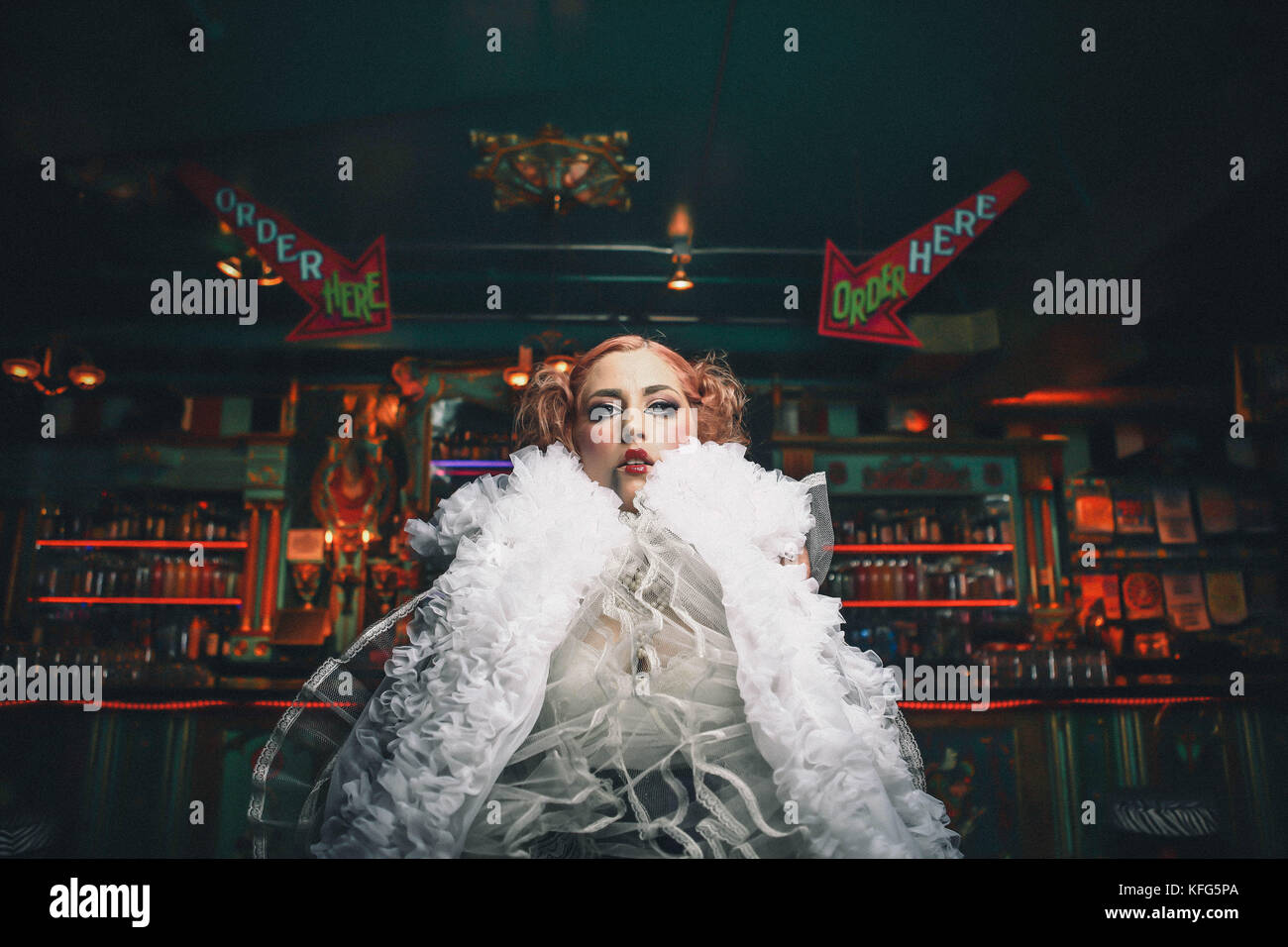 Eccentrico doll face donna con rossetto rosso guance rosee pelle pallida sorge nella parte anteriore del bar indossando volant bianco boa intorno al suo collo pigtail nei suoi capelli Foto Stock
