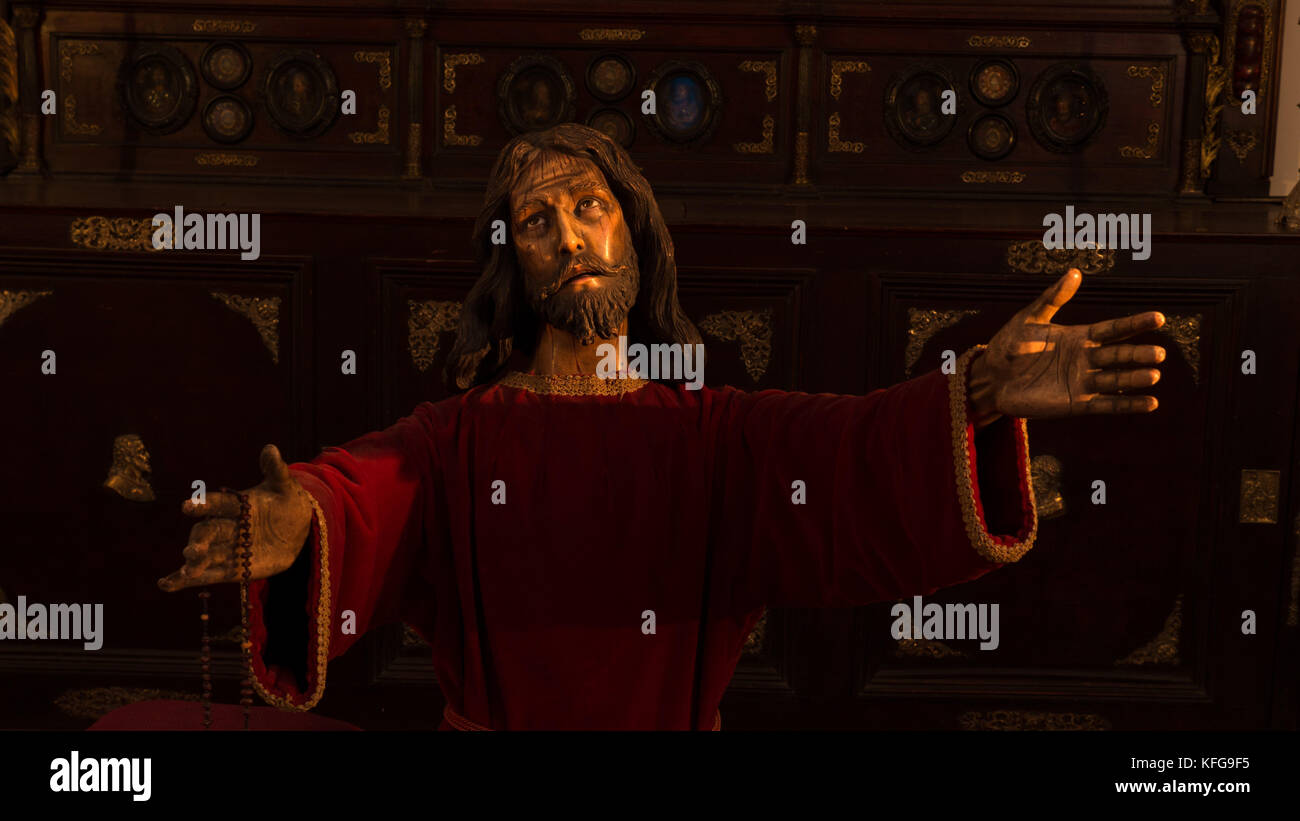 Luce drammaticamente la statua di Gesù Cristo in abito rosso con le mani tese tenendo un rosario con uno sguardo di dolore e angoscia sul suo viso. Foto Stock