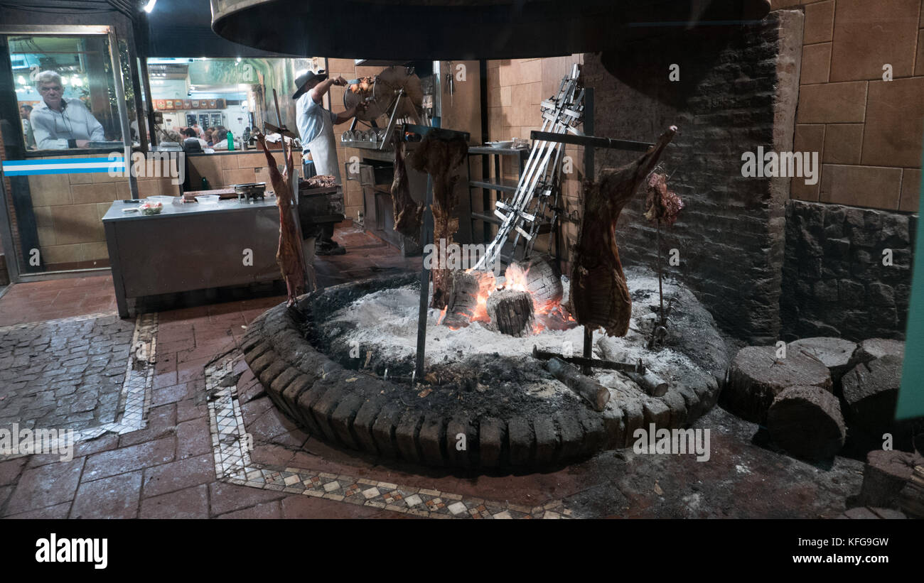 Tradizionali argentini aperto il fuoco di fiamma di cottura delle carni bovine strombato, pezzi di legno in primo piano con lo chef del taglio della carne e il cameriere guardando attraverso la finestra. Foto Stock