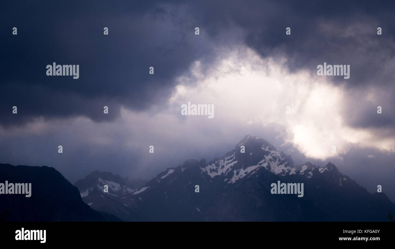 Gli alberi di drammatica del piercing luce attraverso nuvole di tempesta di neve illuminante patagonia rivestito di picchi di montagna al lago Nahuel Huapi National Park in BARILOCHE Foto Stock