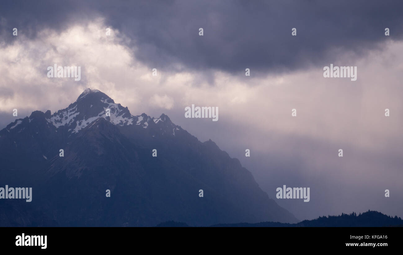 Gli alberi di drammatica del piercing luce attraverso nuvole di tempesta di neve illuminante patagonia rivestito di picchi di montagna al lago Nahuel Huapi National Park in BARILOCHE Foto Stock