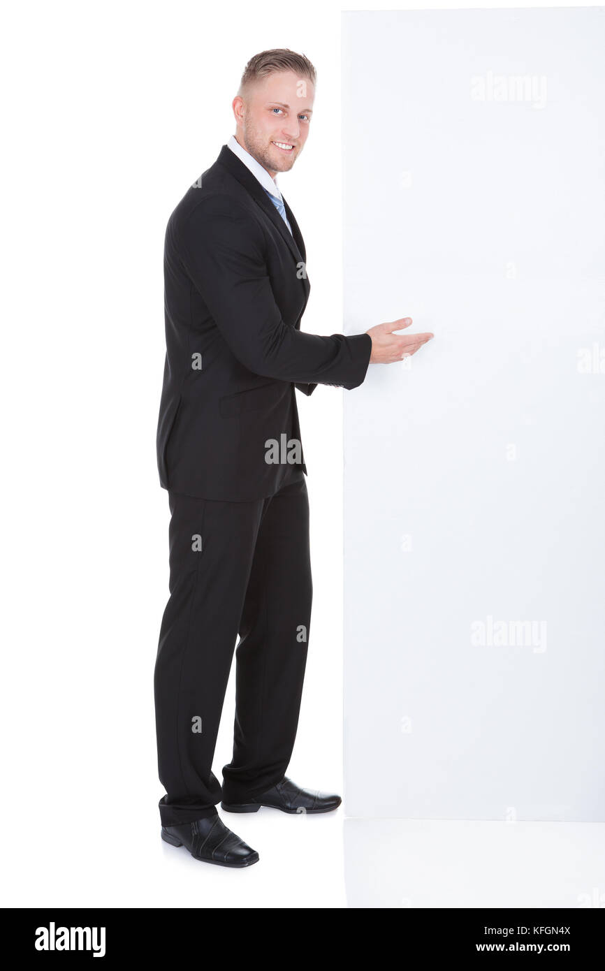 Elegante uomo d affari rivolto in una casella vuota white segno che egli è in piedi accanto a con un sorriso amichevole come egli attira la vostra attenzione al copyspace Foto Stock