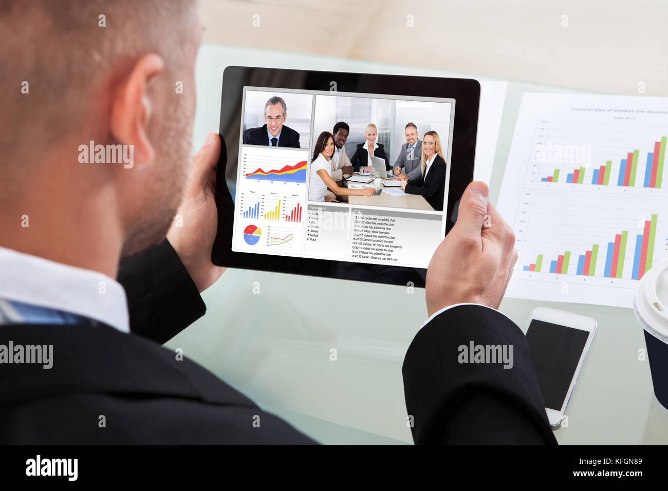 Imprenditore su un video o una chiamata in conferenza su il suo tablet con un'immagine di colleghi di lavoro in occasione di un incontro e grafici a barre sullo schermo Foto Stock