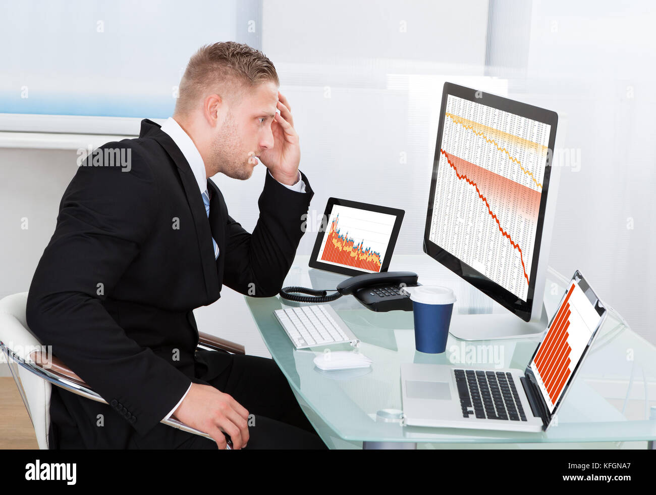 Imprenditore disperato di fronte a perdite finanziarie seduto alla sua scrivania consultazione tre grafici su monitor diversi tutti scenda in rosso Foto Stock