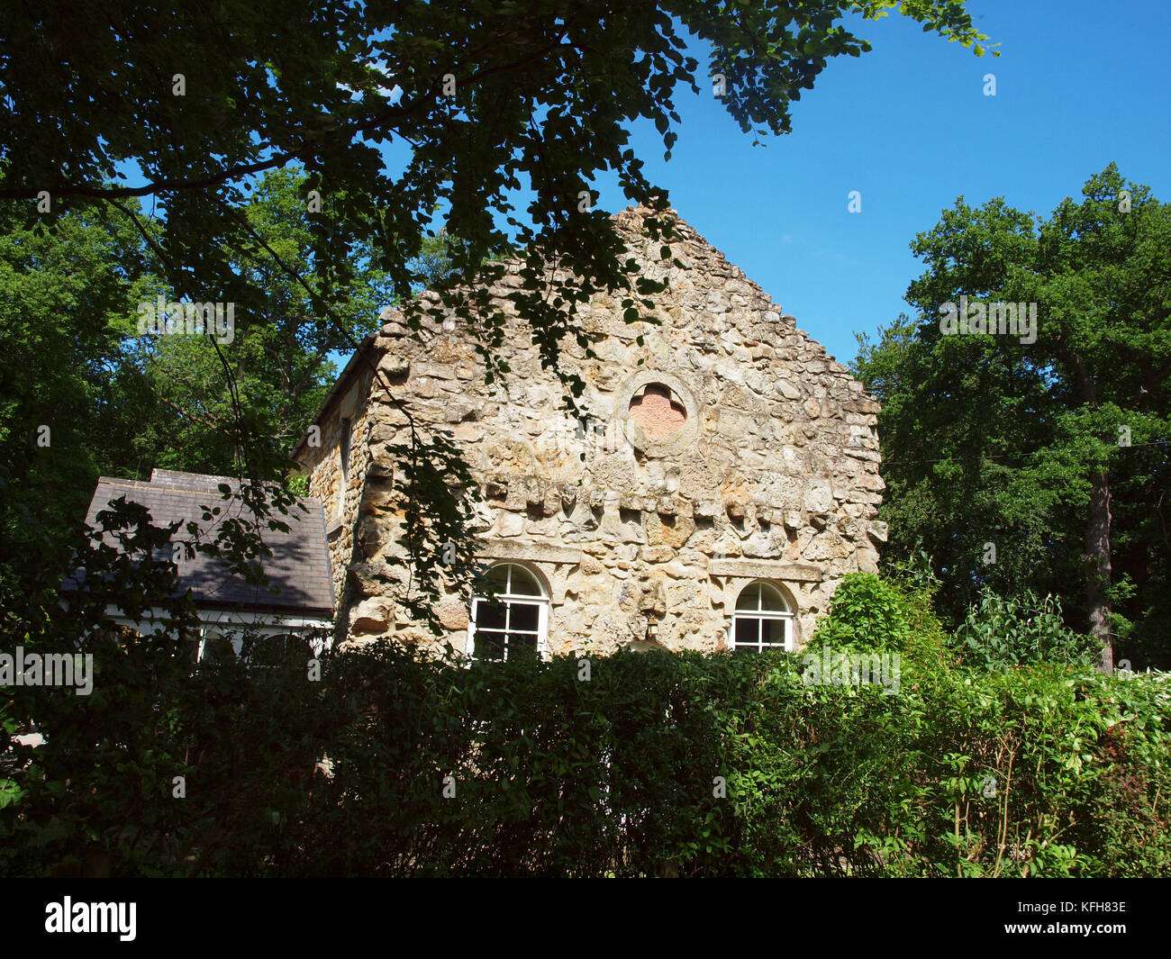 Un cottage con un vecchio muro di pietra facciata situato sulla propria terra tra gli alberi, arbusti e cieli blu. . Foto Stock