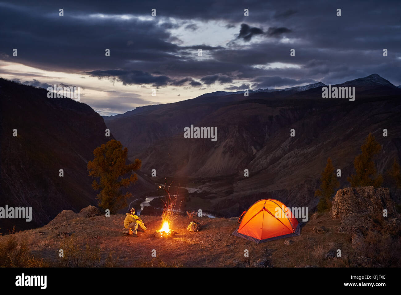 Un uomo vicino alla tenda illuminata e falò in montagna in Alba Foto Stock