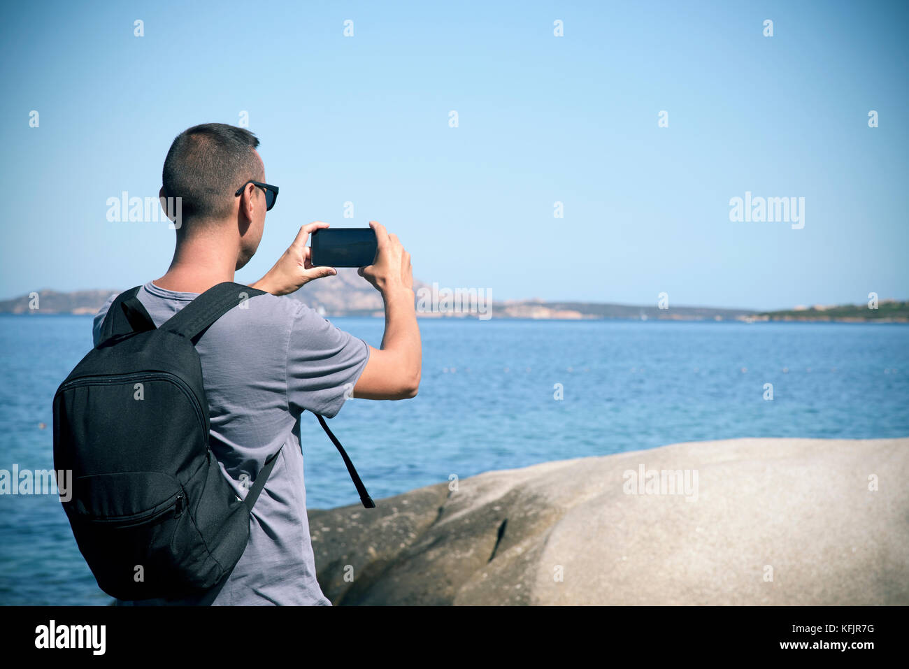 Primo piano di un giovane caucasico con uno zaino visto da dietro scattando una foto, con il suo smartphone, del Mar Mediterraneo a Cala Ginepro beac Foto Stock