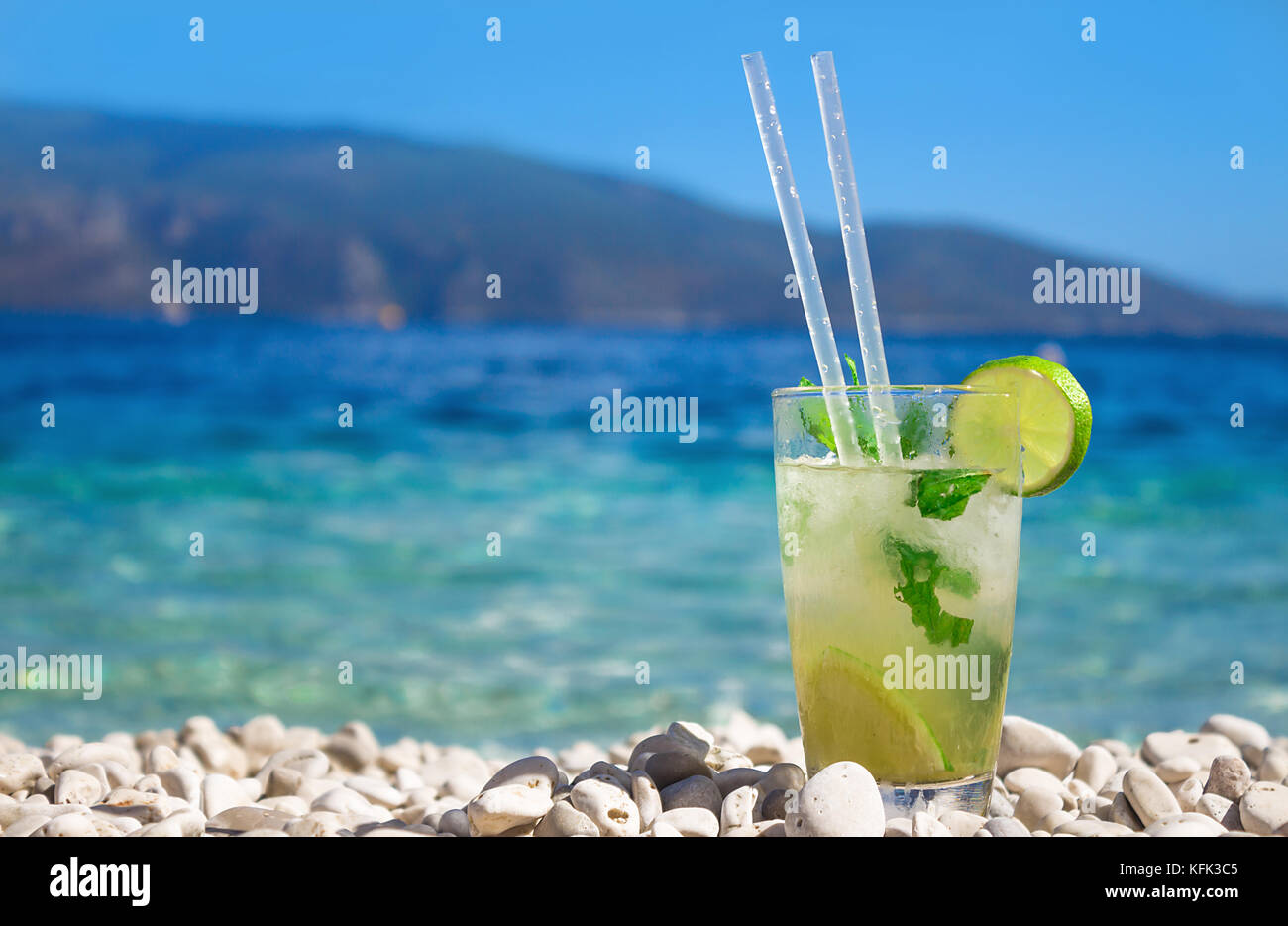 Bicchiere di freschi cocktail mojito con ghiaccio, lime e menta sulla bianca spiaggia di ciottoli sul mare turchese su vacanze estive presso l'isola di Cefalonia, Grecia Foto Stock