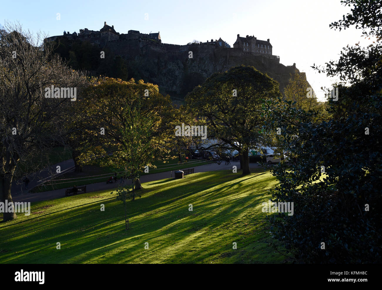 La luce solare bassa getta ombre attraverso gli alberi nei giardini di Princes street, con il Castello di Edimburgo stagliano in background. Foto Stock