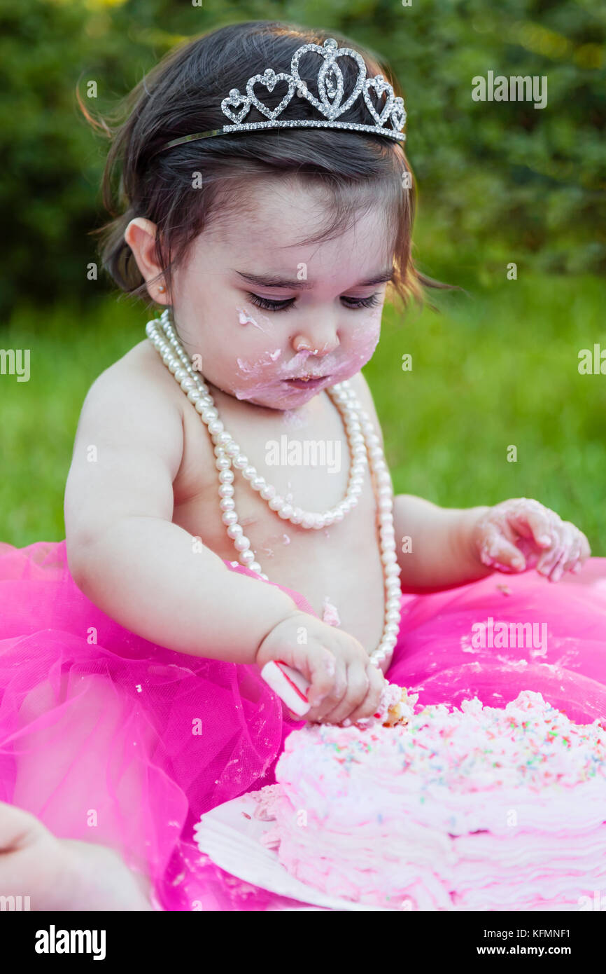 Happy baby toddler girl in primo compleanno festa anniversario, con la faccia sporca facendo un pasticcio di torta rosa con candela. princess tiara, perle e tutu Foto Stock