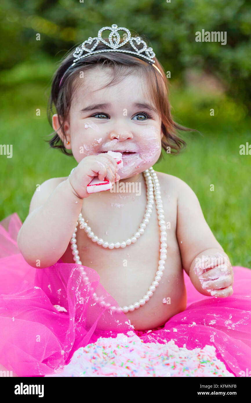 Sorridente happy baby toddler girl celebrando primo compleanno festa anniversario. holding e masticare candela con la faccia sporca da rosa torta, facendo un pasticcio Foto Stock