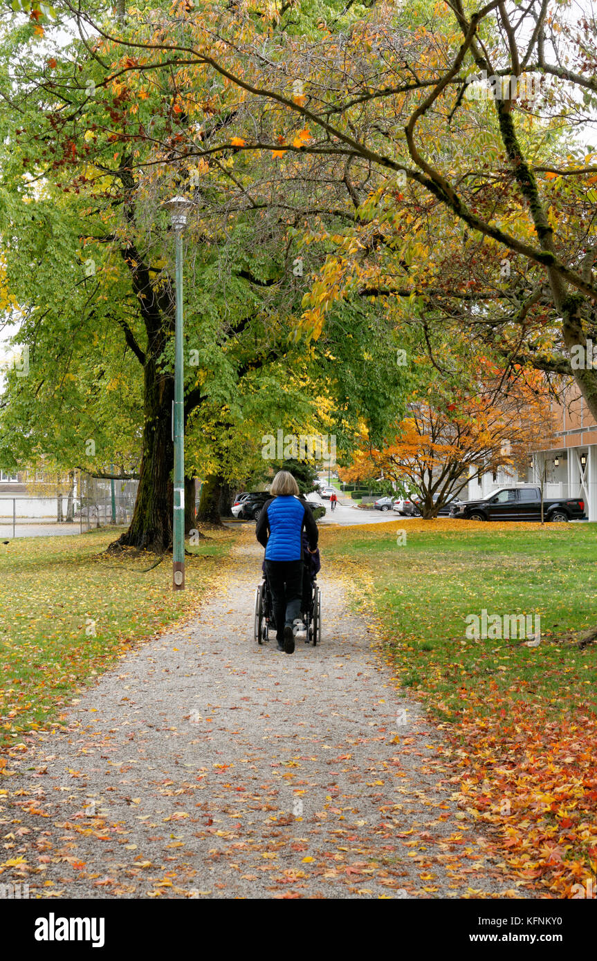 Mddle in età donna spingendo una persona anziana in una sedia a rotelle attraverso un parco in autunno, Vancouver, BC, Canada Foto Stock