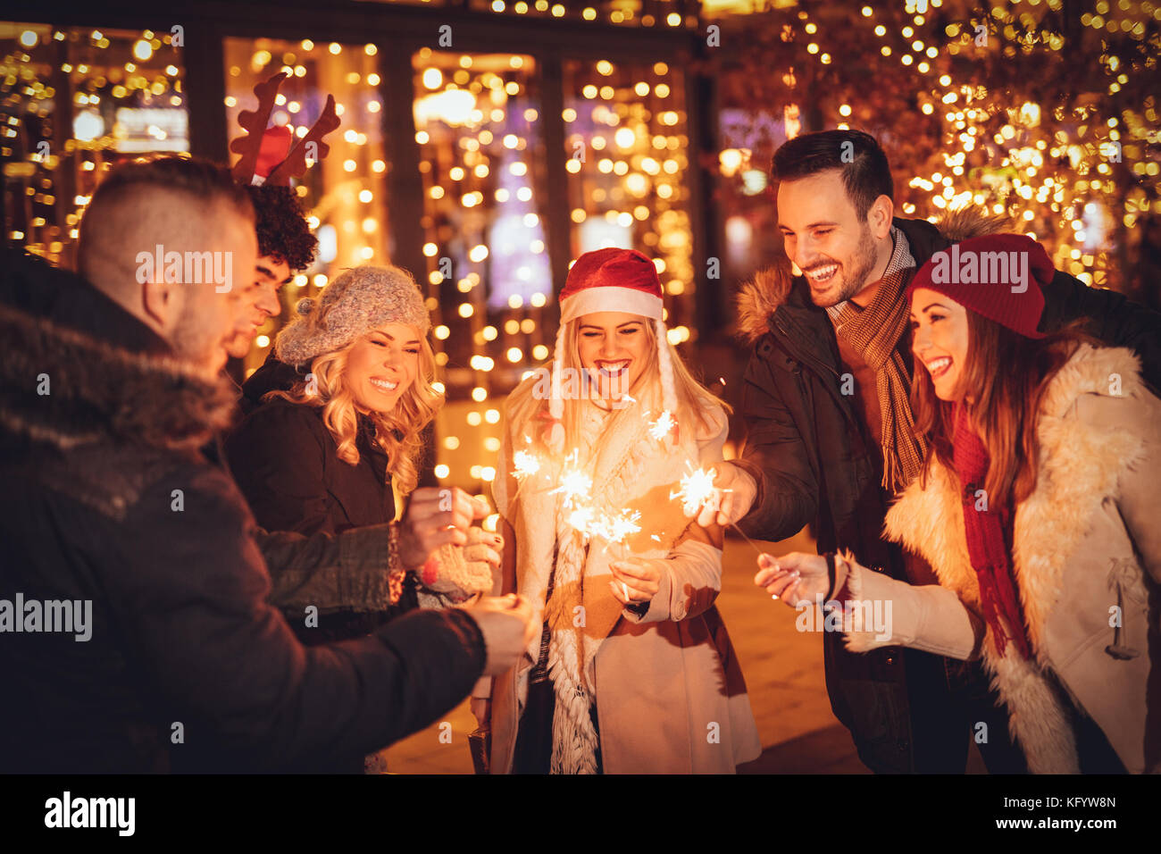 Un gruppo di giovani amici di capodanno con bastoncini di scintilla divertirsi nella città Street di fronte luminosa finestra del negozio. Foto Stock