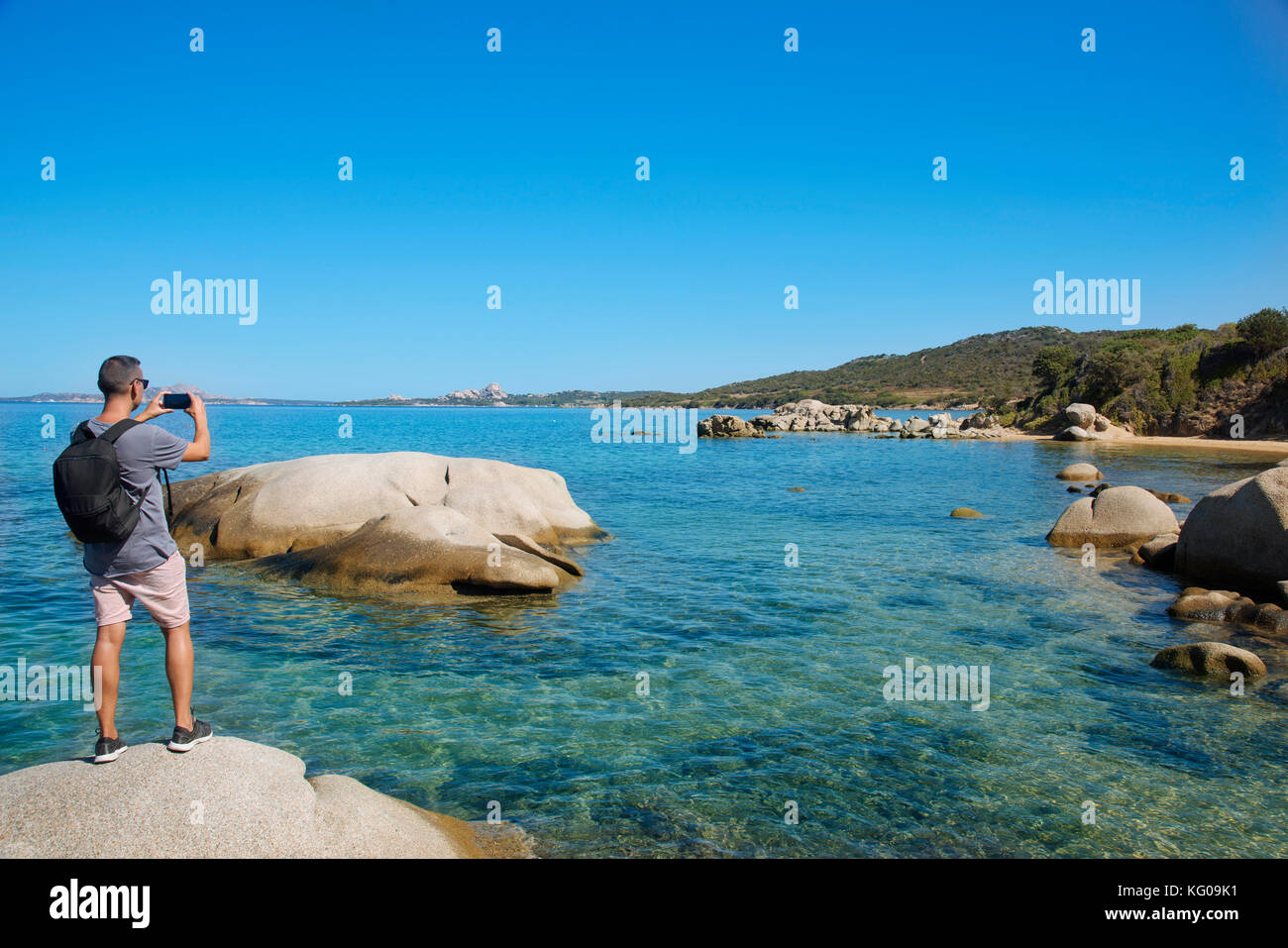 Un giovane caucasico con uno zaino visto da dietro scattando una foto, con il suo smartphone, del Mar Mediterraneo alla spiaggia di Cala Ginepro, nella C Foto Stock