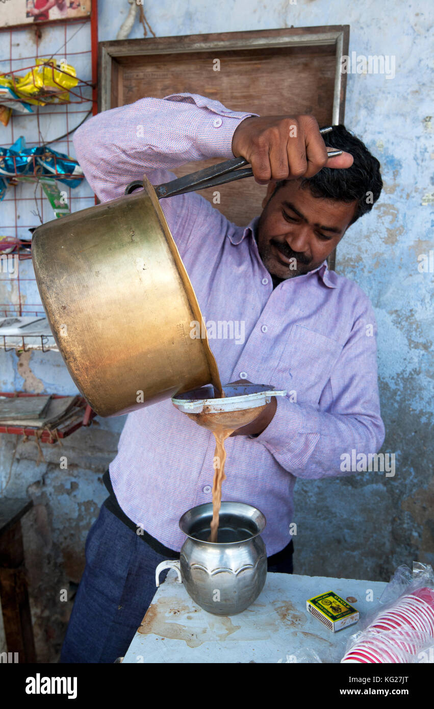 Chai wallah versando chai dalla coppa di ottone, servendosi di un paio di pinze, attraverso il crivello inferiore nel recipiente stagno, in diggi Village market, diggi, Rajasthan, India, Asia Foto Stock