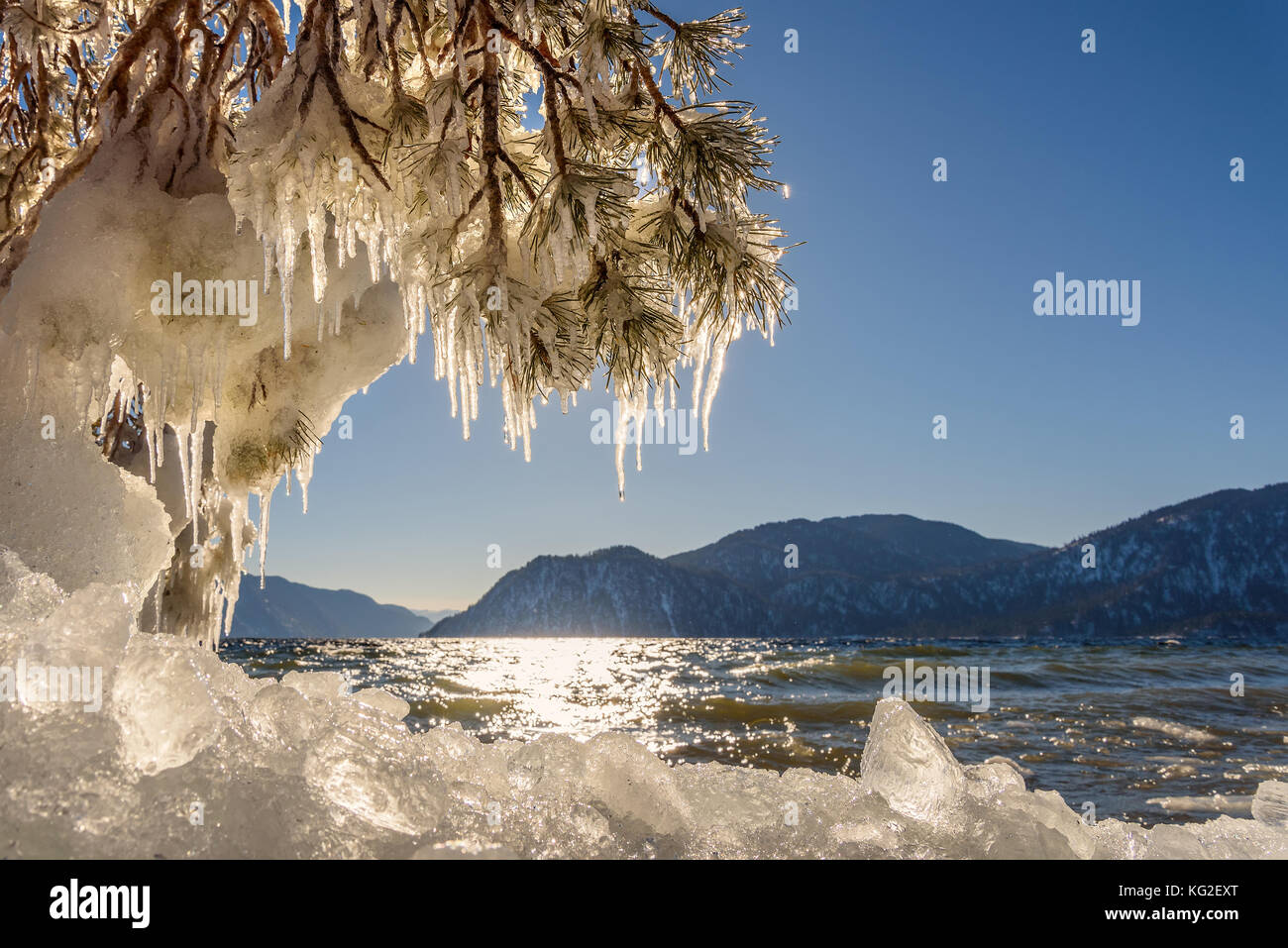Bellissimi paesaggi invernali con il lago e le montagne di ghiaccio, sulla riva, i riflessi del sole sull'acqua e rami di pino con ghiaccioli in una giornata di sole Foto Stock