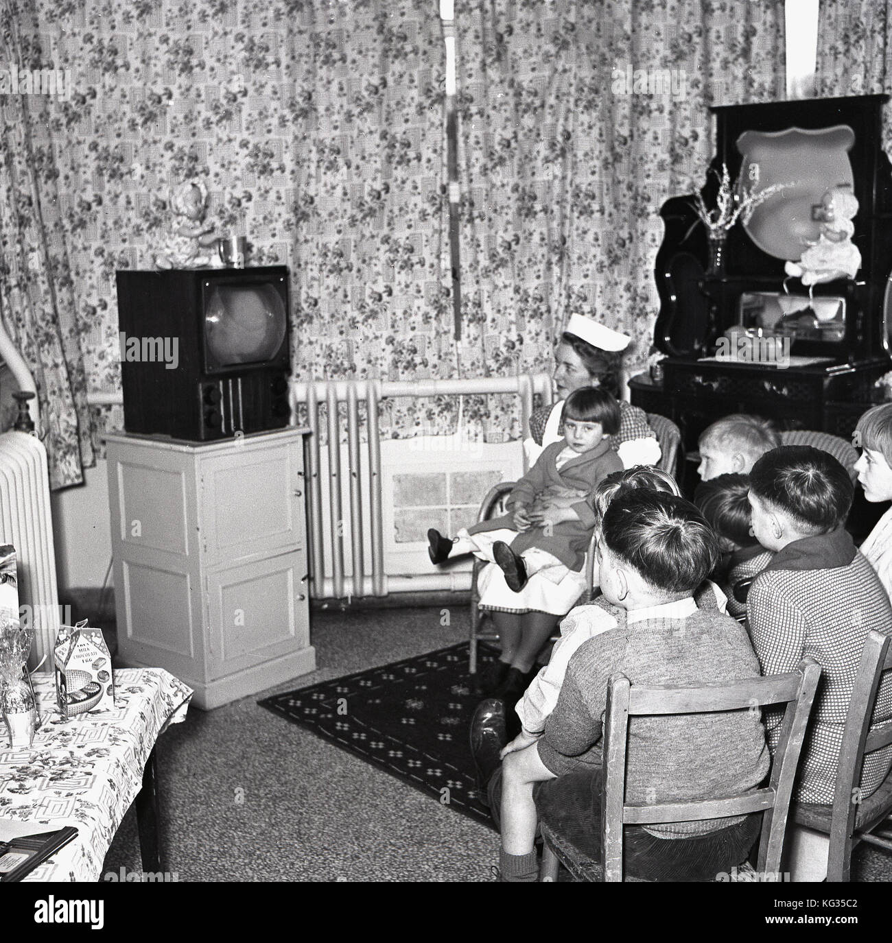 Degli anni Cinquanta, Inghilterra, sala TV in un ospedale per bambini, gruppo di piccoli bambini seduti insieme a guardare la televisione con una giovane ragazza detenute da un infermiere. Foto Stock