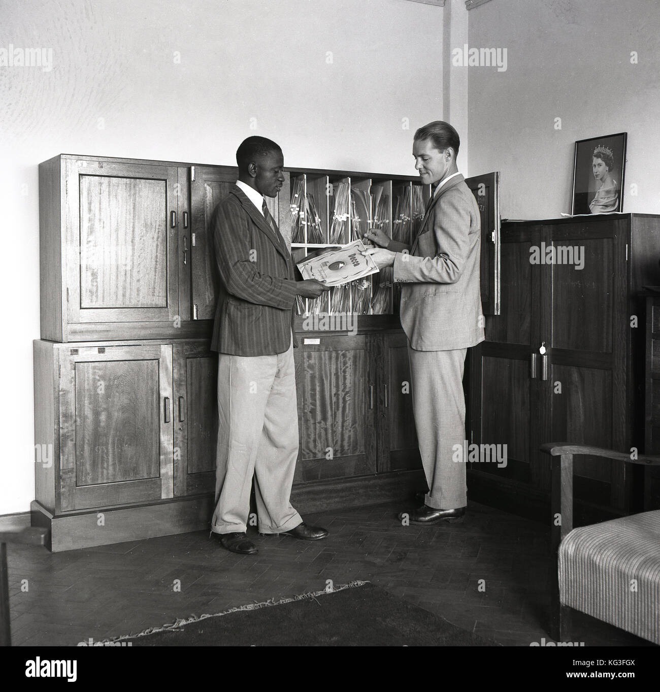 Degli anni Cinquanta, foto storiche che mostra un uomo britannico in tuta presentando un lungo riproduzione dischi in vinile,un Decca LP, ad un visitatore d'oltremare, Inghilterra, Regno Unito. Una cornice per una foto di una giovane regina Elisabetta è seduta su di un armadio. Foto Stock