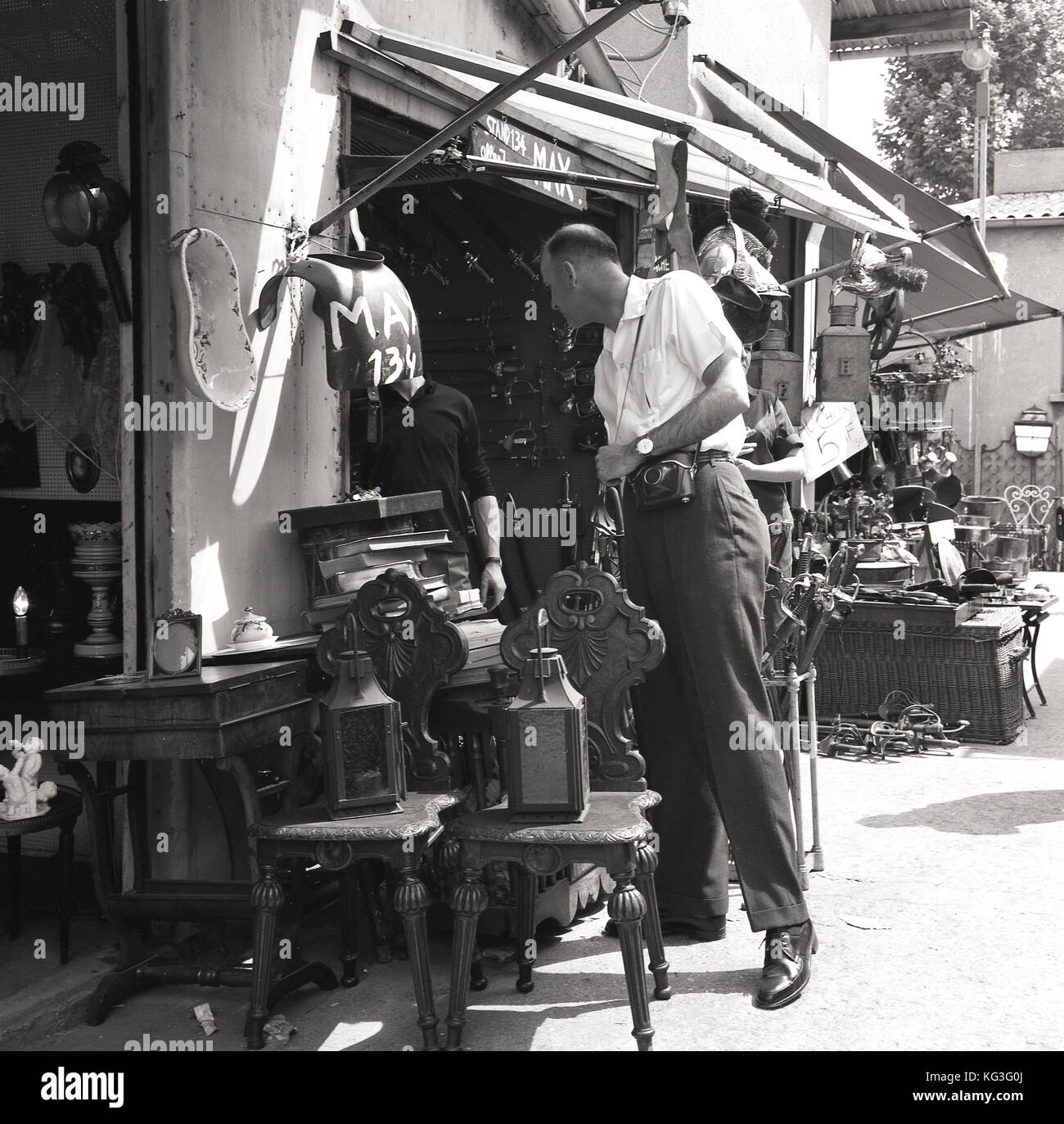 1960s, storico, un turista maschile con macchina fotografica dora una stalla piena di mobili, grès e altri oggetti d'antiquariato al famoso mercato delle pulci Les Puces a Parigi. Foto Stock
