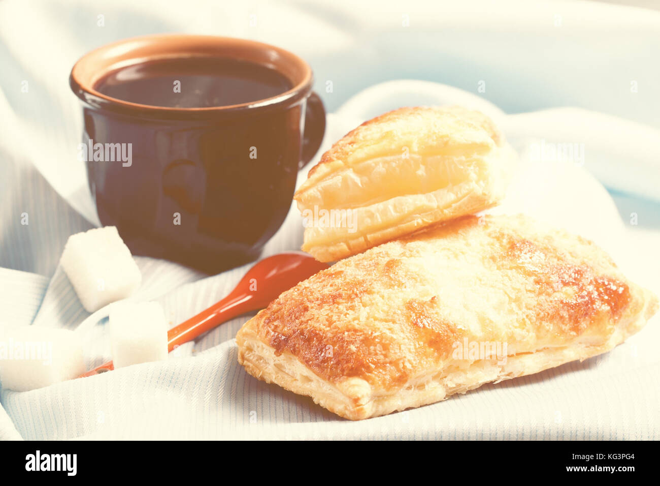 Caffè e panini freschi al sole del mattino. una coppa in ceramica da caffè e un rotolo di pasta sfoglia su un tovagliolo. Vicino a cucchiaio e pezzi di zucchero. è Foto Stock