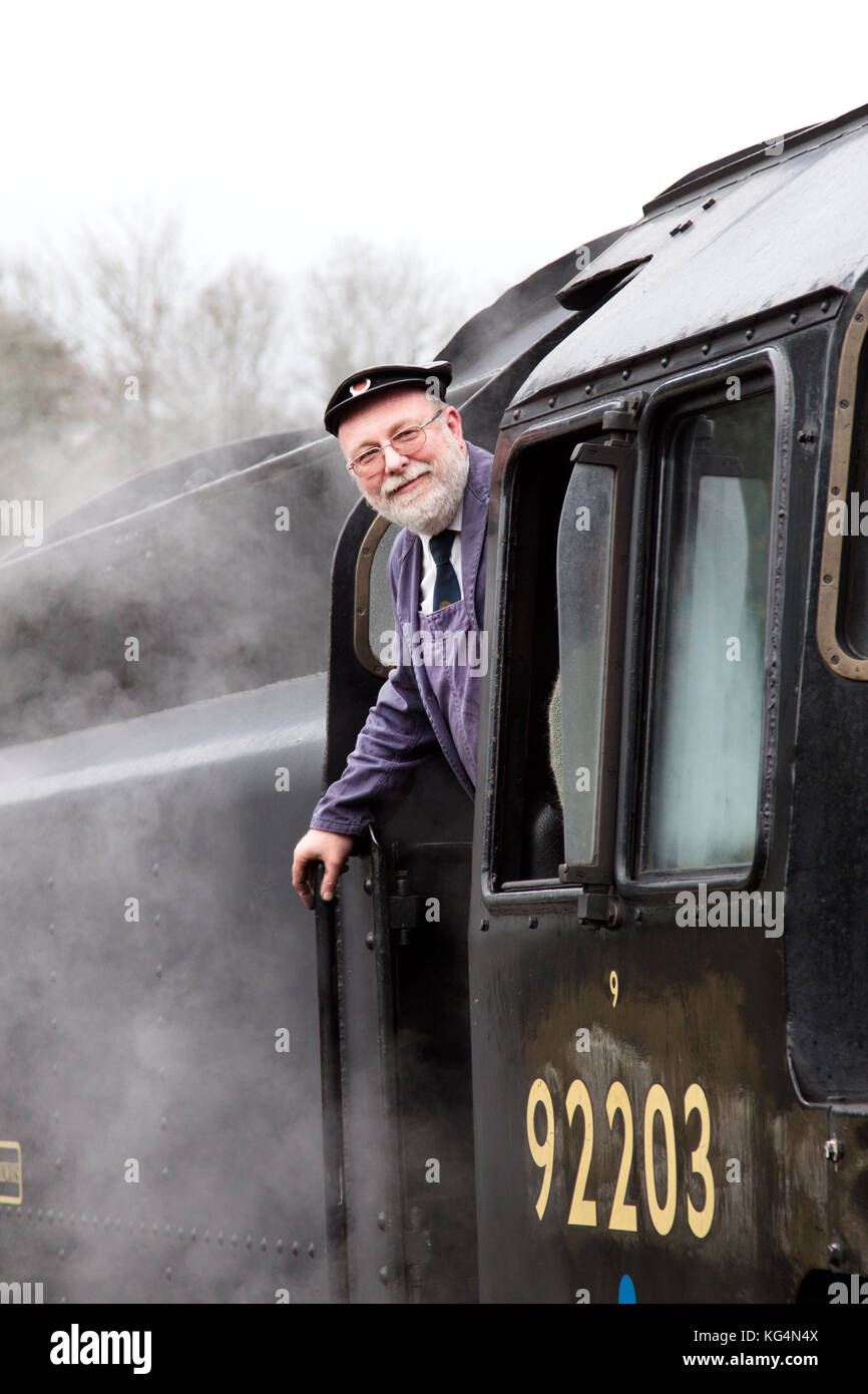 Un conducente si appoggia all'esterno della cabina con un getto di vapore treno che è temporaneamente fermata ad una stazione ferroviaria Foto Stock