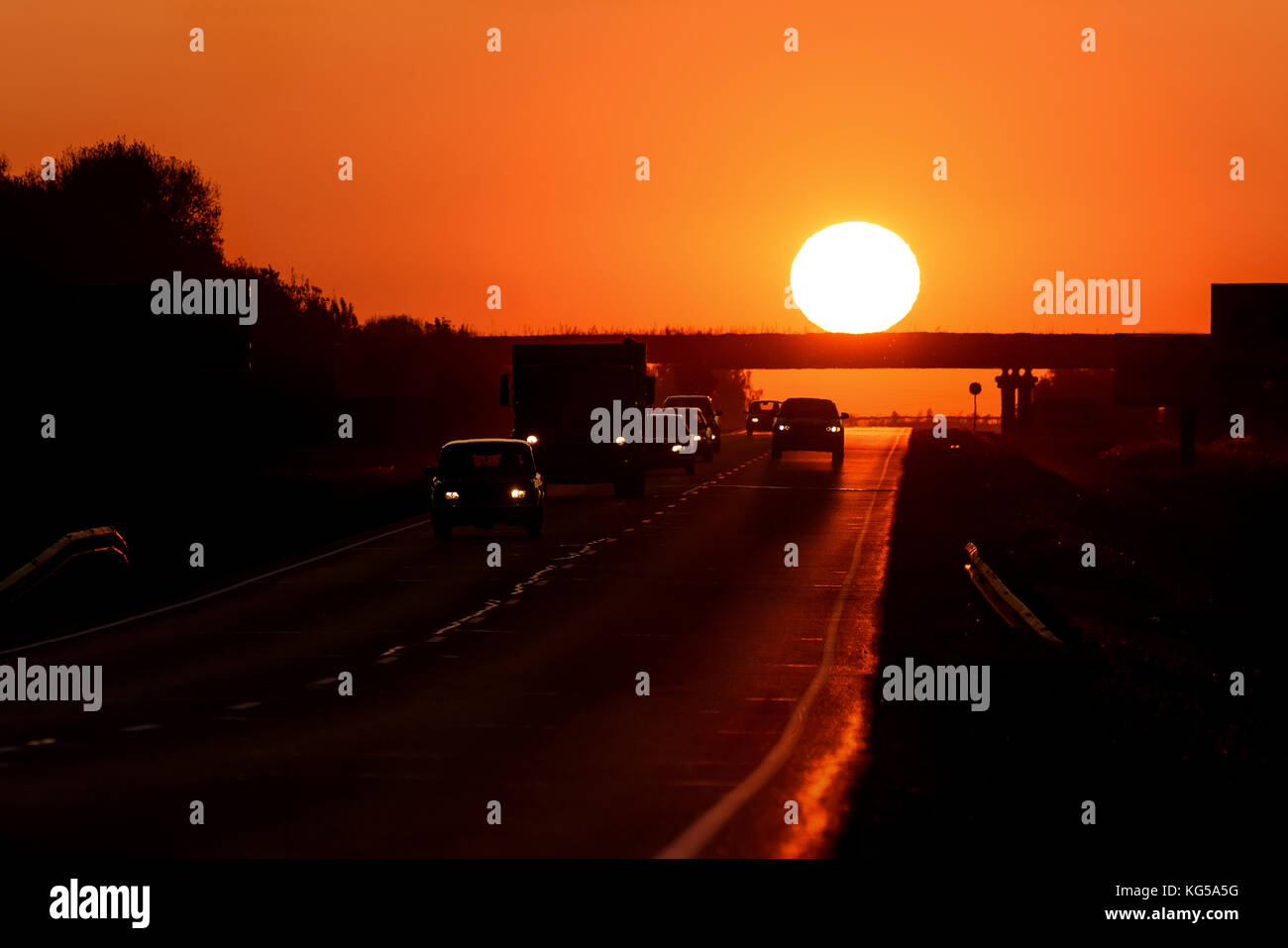 Colorato sfondo astratto con una strada asfaltata, automobili e un sole rotonda su un ponte contro lo sfondo di un rosso infuocato-cielo arancione al tramonto Foto Stock