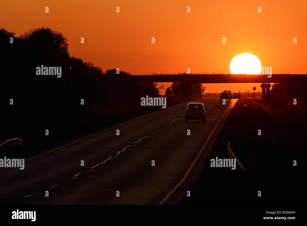 Colorato sfondo astratto con una strada asfaltata, automobili e un sole rotonda su un ponte contro lo sfondo di un rosso infuocato-cielo arancione al tramonto Foto Stock
