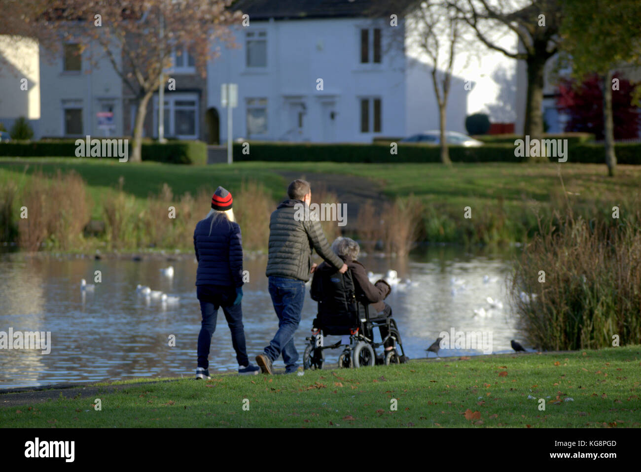 Coppia giovane spingendo invalida le donne anziane in sedia a rotelle nel parco accanto al laghetto visto da dietro Foto Stock