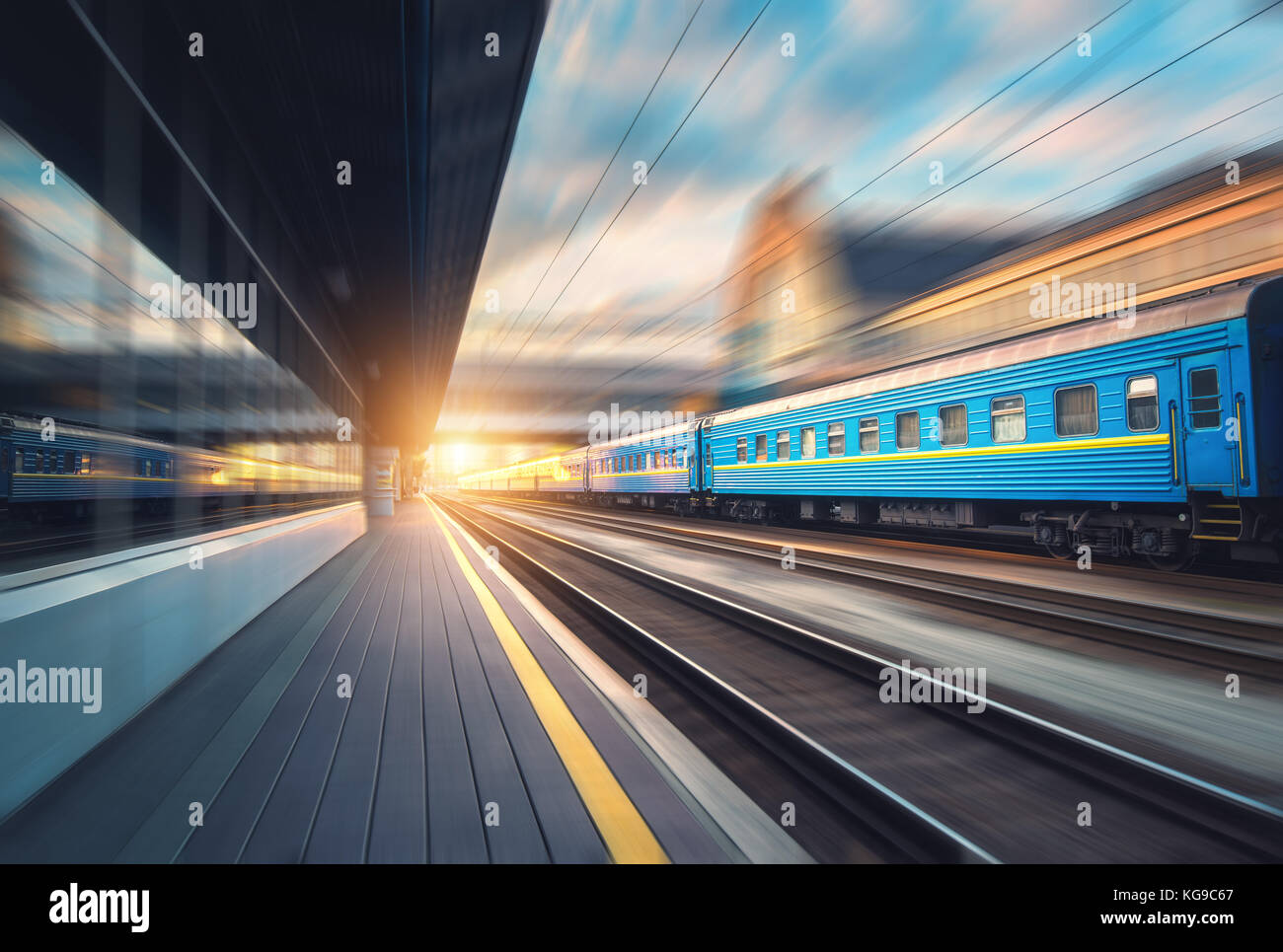 Bellissimo il treno blu con i carri in movimento con la stazione ferroviaria al tramonto. visione industriale con il moderno treno, ferrovia, piattaforma ferroviaria, edifici, Foto Stock